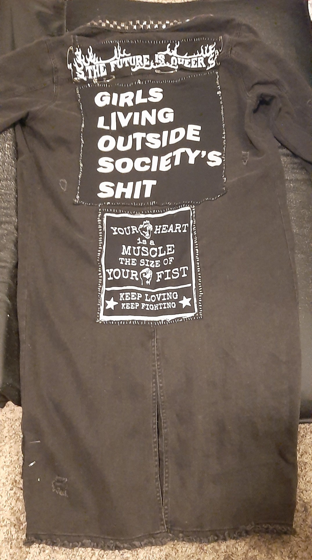 `the back of my black denim trenchcoat. it has a big patch that says GIRLS LIVING OUTSIDE SOCIETY'S SHIT and the future is queer and your heart is a muscle the size of your fist
