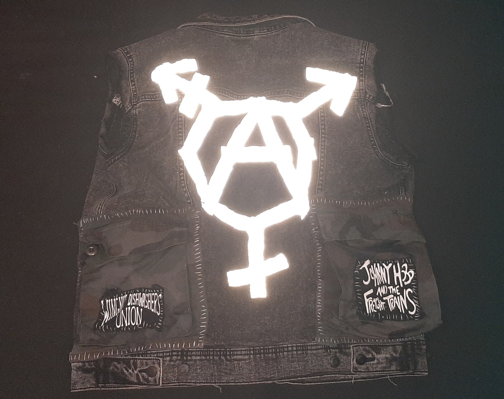 back of my grayish black denim punk vest. it has two large military pockets sewn on with johnny hobo and wingnut dishwashers union patches atop them. there's a trans anarchy symbol made in reflective tape, lit bright by the flash
