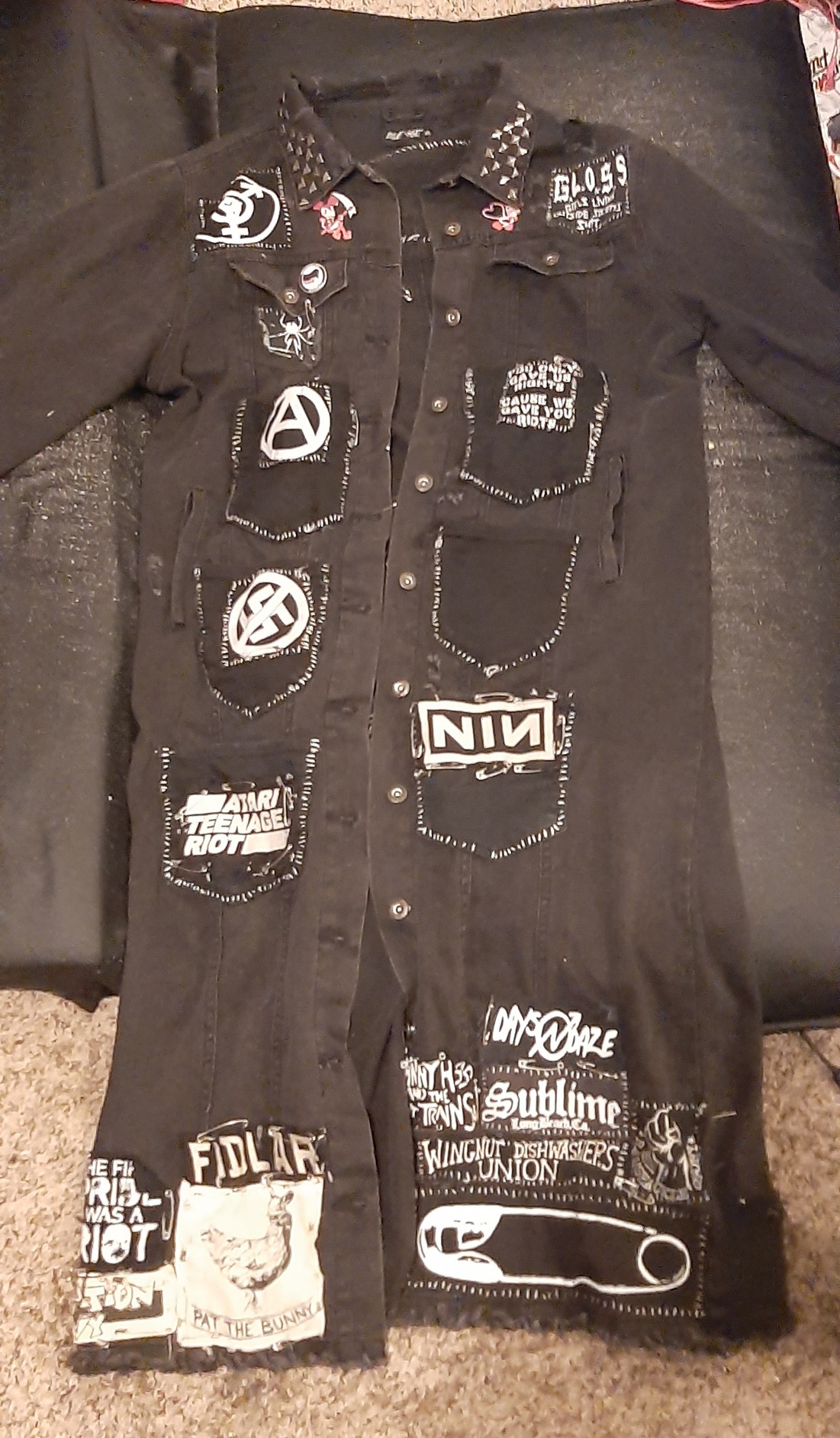 the front of my black denim trenchcoat covered in various pins, studs, and patches. there are six black denim pockets sewn onto the front